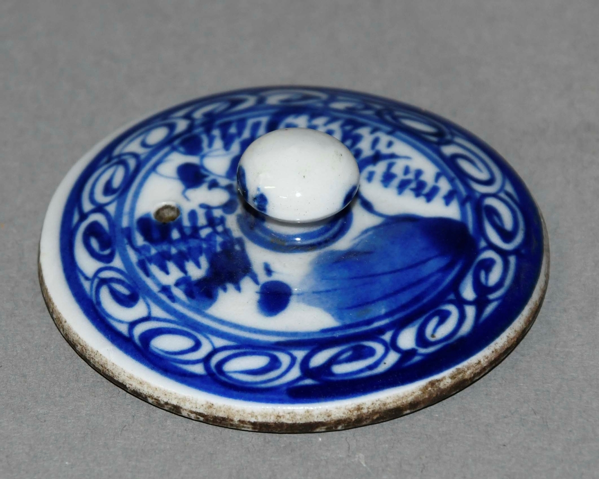 Lokk til tepotte av hvit keramikk. Lokket er rundt og har en knott på midten. Lokket har blå dekormaling.