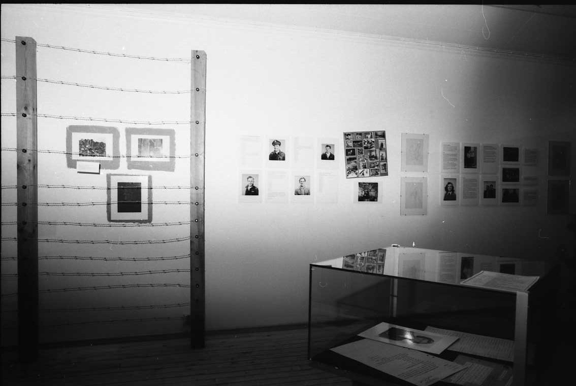 Dokumentation av utställning: G H Trapp och tre sekler av främlingshistoria. Stolpar med taggtråd framför vägg med text och bild.