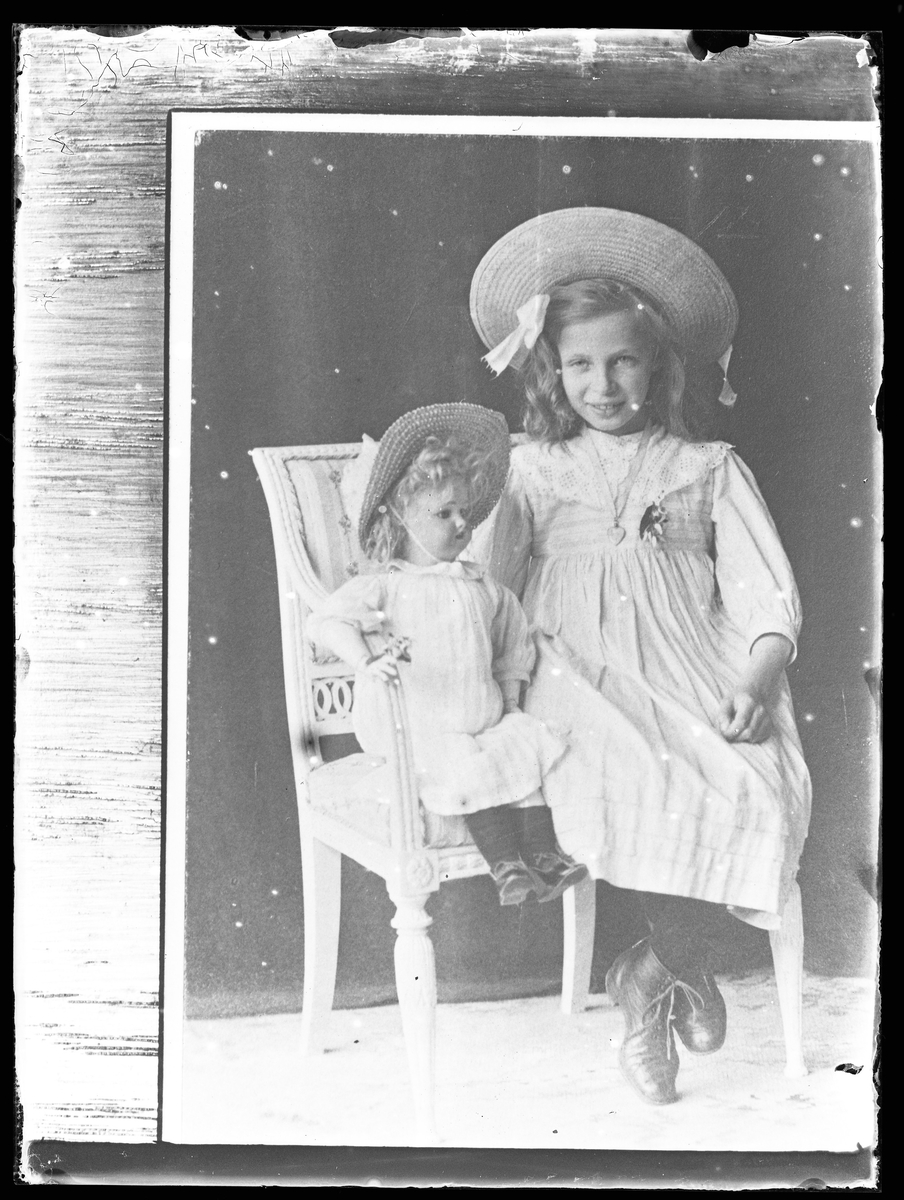 Reprofotografi på beställning av fru Lenander (tolkat som Anna Lenander). Bilden är ett porträtt av en flicka i ljus klänning och halmhatt som sitter på en stol tillsammans med en porslinsdocka.