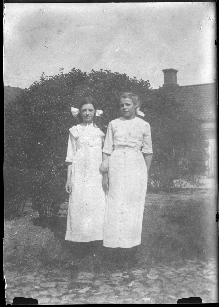 Reprofotografi av bild på två flickor, klädda i vita klänningar och med rosetter i håret, fotograferadde framför en buske. I bakgrunden skymtar hus. I fotografens anteckningar står "Rep. för Dahlgren"