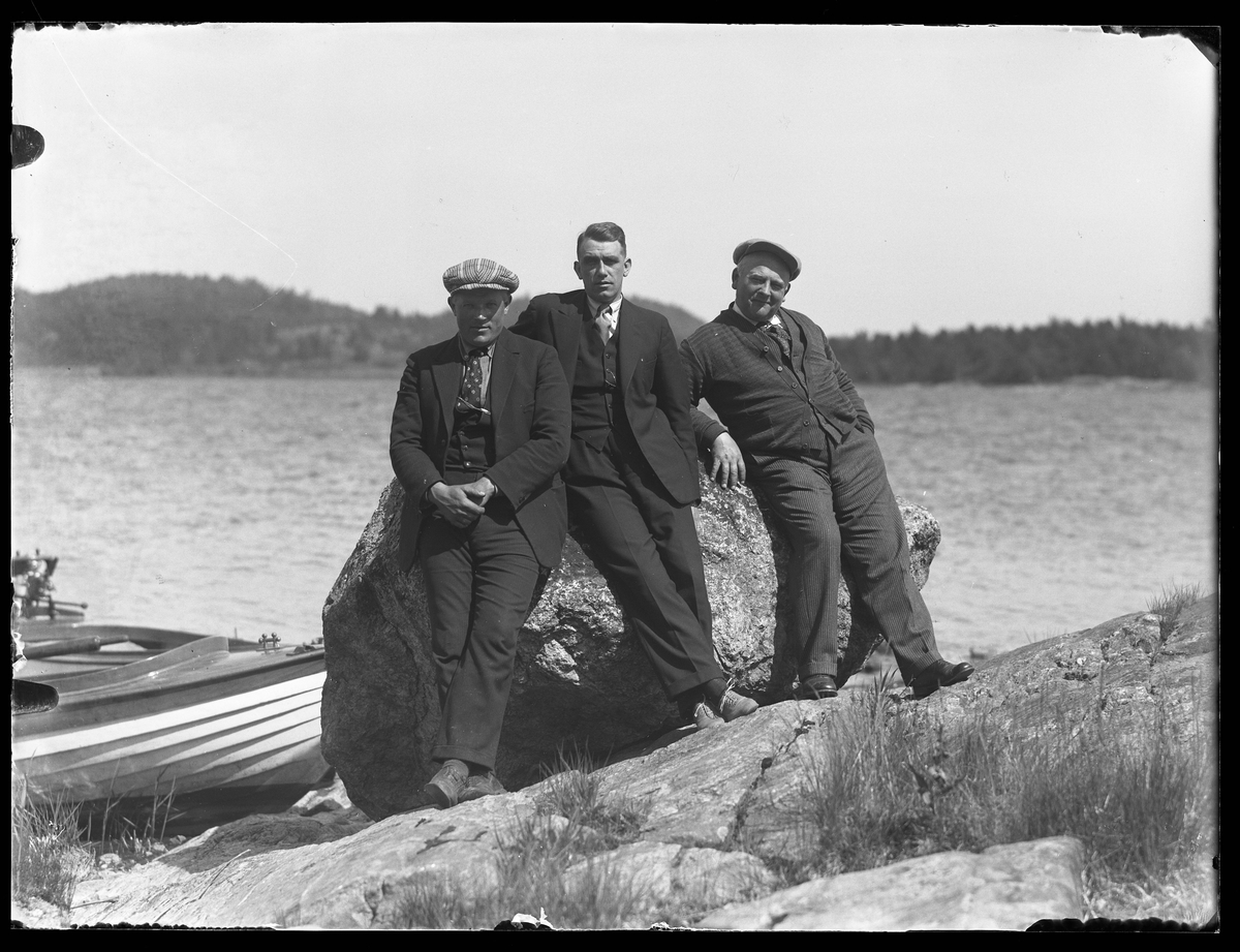 Tre mån står lutade mot en sten på en klippa vid en sjö. Bakom dem ligger en båt förtöjd. I fotografens egna anteckningar står "Olle, Andersson, Sandin, Kvarnholmen"