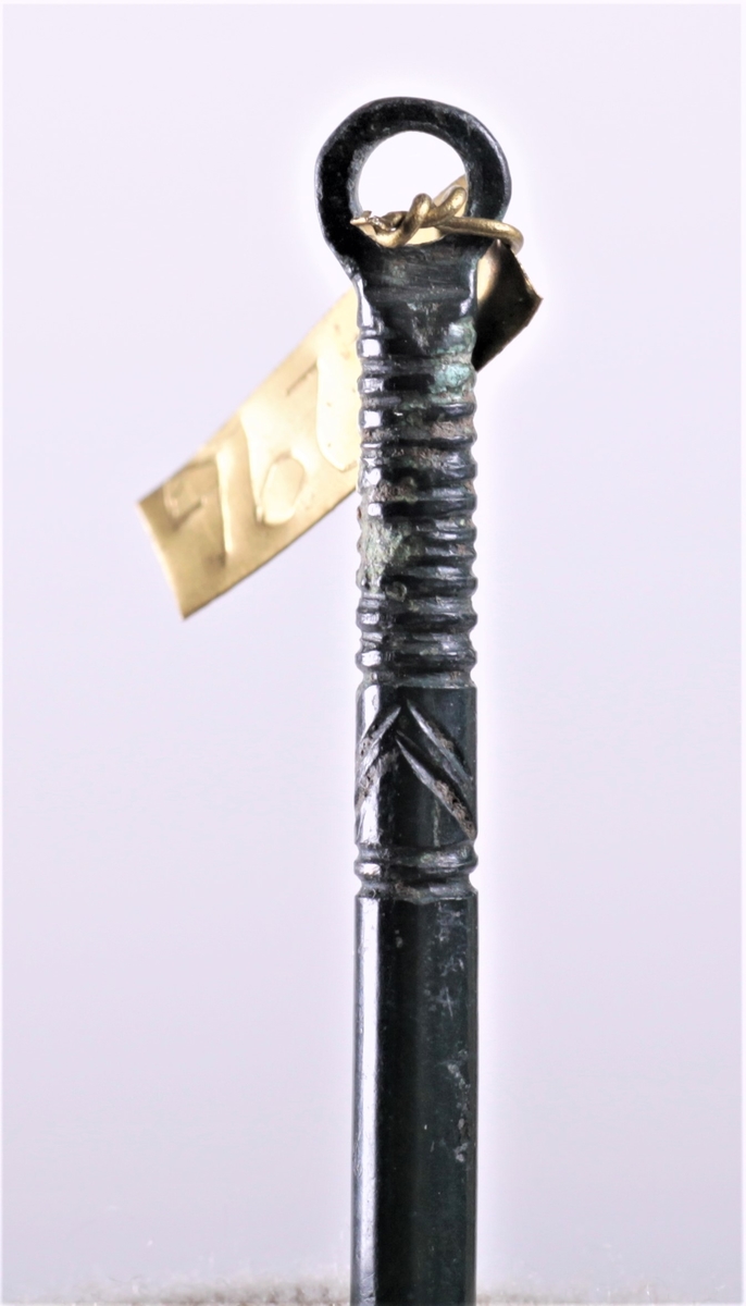 Smykkenål i bronse funnet i en tidligre gravhaug. En lang, spiss nål med et rundt øye. Nålen er dekket av sort emalje som kalles niello.