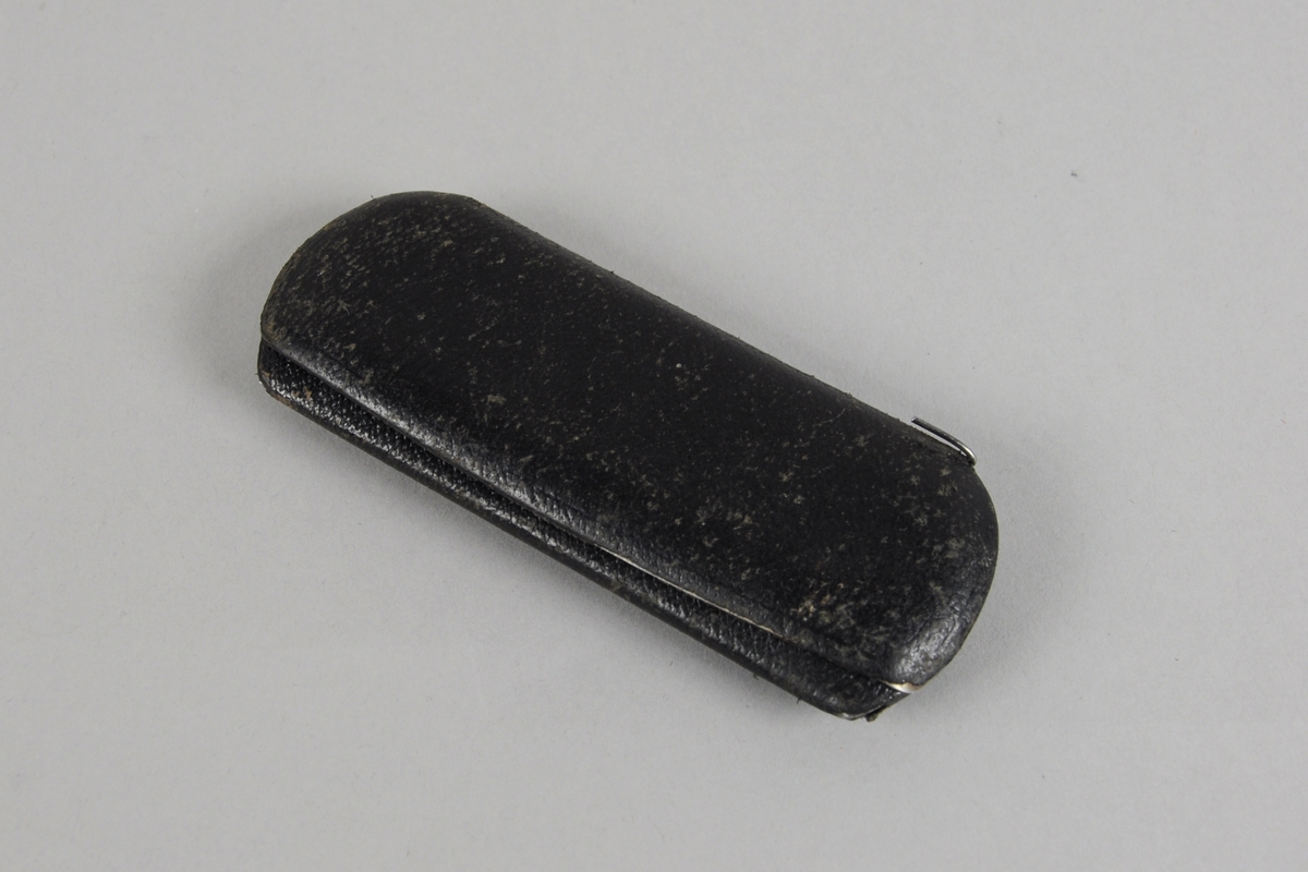 Brilleetui av metall med svart trekk, som åpnes på langsiden. Svart fór. I etuiet er det en lorgnett og en pusseduk.