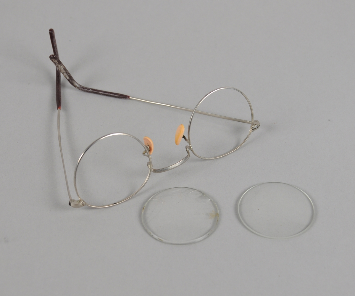 Brillerinnfatning av metall med plast ytterst på brillestengene. Innfatningen ligger i et etui, der det også ligger et par runde brilleglass som ikke passer til innfatningen.