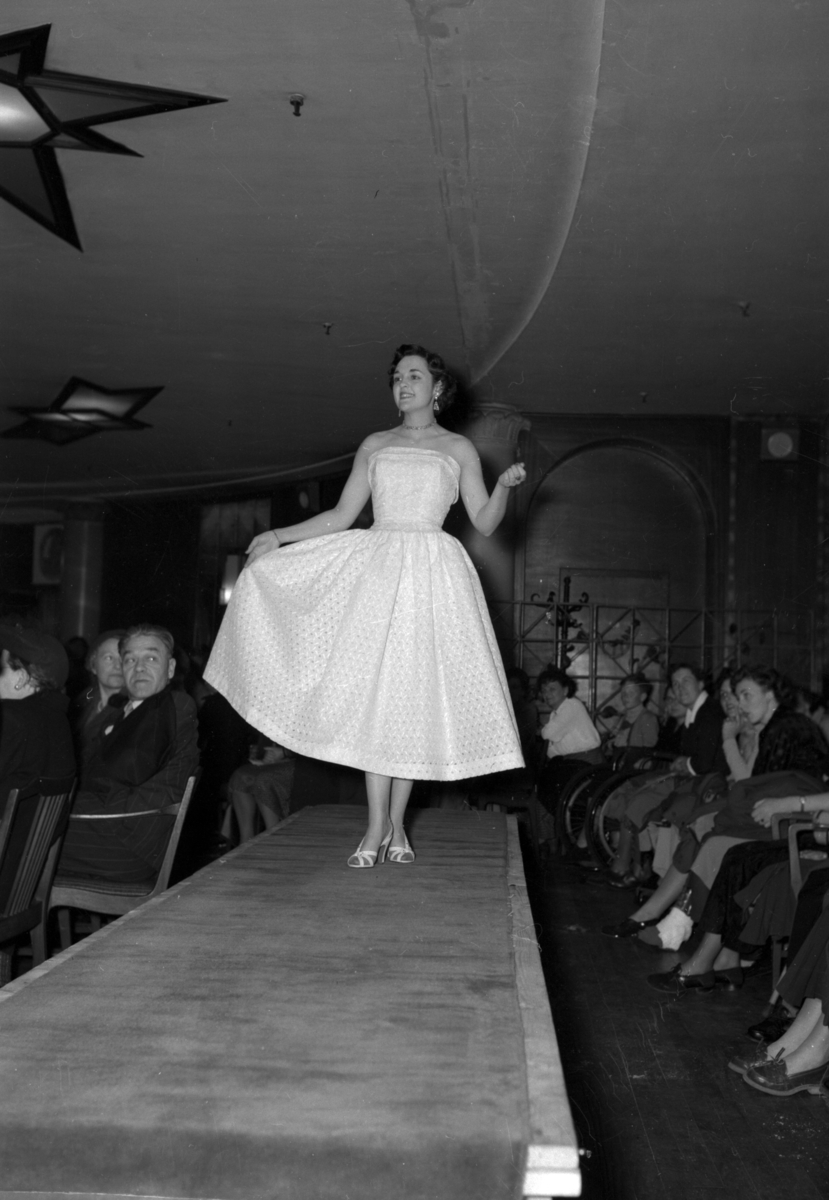 En kvinne på podiet iført kjole uten stropper. Fra en motevisning på Steen & Strøm fra 7. mai 1954..