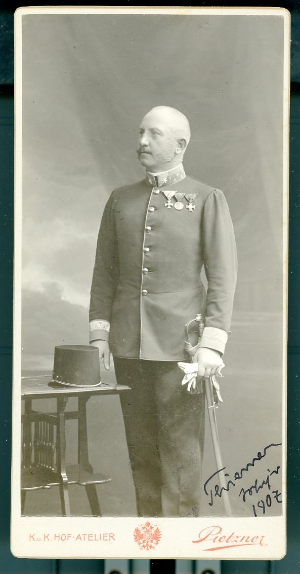 Kabinettsfotografi: Österrikisk-ungersk överste med medaljer på bröstet. Uniformsmössan liggande på ett bord och handen vita handskar på värjan. Hans namn är svårt att tyda.