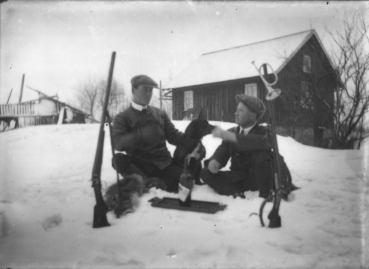 Två män med jaktbyte och gevär samt ett jakthorn vintertid.