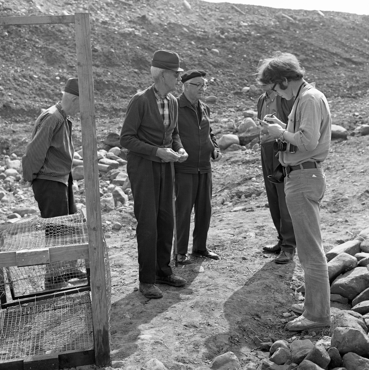 Til høyre på dette bildet ser vi Åsmund Eknæs (1940-1981) på feltarbeid på Fossbakken under garden Nedre Kåterud i Stange, der det tradisjonelle kupefisket etter mort (her kalt «sørenne») i den nedre delen av Svartelva, som på dette tidspunktet var truet av en utvidelse av hovedvegen mellom Oslo og Trondheim, E6-  Eknæs er fotografert med en notatblokk i hendene og kamera på magen mens han snakket med fire informanter, blant annet Martinus Hovie (lengst til venstre), Håkon Berge og Birger Børresen (skjult bak Eknæs).  Mannen som sto som nummer tre fra venstre har vi foreløpig ikke navnet på. Til venstre i bildet ser vi ei av kupene (rusene).  Mens de tradisjonelle kupene i dette fisket var lagd av trespiler med bomullsnett, var de som ble brukt de siste åra øyensynlig lagd av nettingtyper som ellers ble brukt i hønsehus og pelsdyrfarmer.

Åsmund Eknæs var fra Krokstadelva i Nedre Eiker i Buskerud.  Han avla magsistergradseksamen i etnologi ved Universitetet i Oslo med avhandlingsarbeidet «Det tradisjonelle laksefiskeriet i Drammenselva - Naturforhold og redskapsutvikling» i 1972  Deretter ble Eknæs ansatt som vitenskapelig assistent ved Norsk Skogbruksmuseum i Elverum.  I 1974 fikk han konservatortittel, etter at en vitenskapelig komité hadde gjennomgått den faglitterære produksjonen hans.  Eknæs var sentral da Norsk Skogbruksmuseum bygde basisutstillinger om ferskvannsfiske, jaktvåpen og tollekniver, og han var en flittig feltarbeider, særlig på steder der det pågikk fiskeaktiviteter.  I 1979 gav han ut boka «Innlandsfiske», som gir en oversikt over innlandsfiskets historie her til lands.  Han foreleste også for grunnfagsstudentene i etnologi om dette emnet.  Eknæs ble dessverre rammet av kreft og døde i 1981, bare drøyt 40 år gammel.
