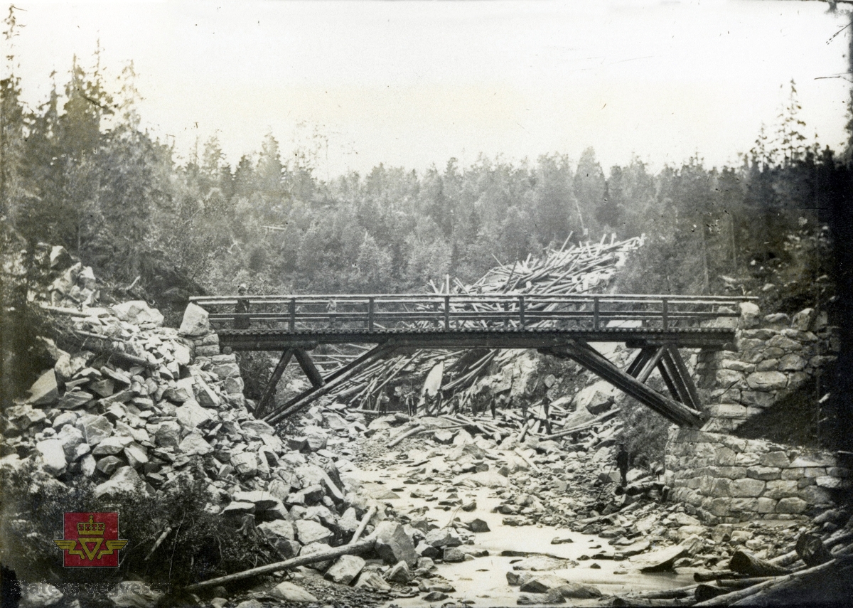 Fiskløselv bru. En person på broen, 12 herrer står ved tømmeret  i bakgrunnen i elveleiet. Et av de første foto fra bruer i fylket.