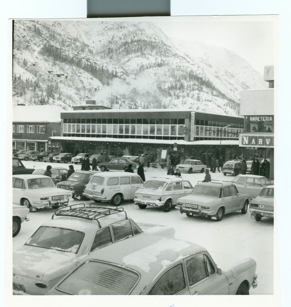 Torvet i Mosjøen 11.mars 1971. Biler er parkert både på torvet og i Strandgata. Jernbanestasjonen. Aluminiumsverket.