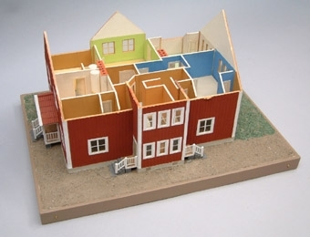 Modell i skala 1:50  av personalbostad, falurött med sadeltak i tegel med vita fönster och skorstenar.
Förstukvisten är valmad med två ingångar via trappor. Innehåller två lägenheter 
om 4 rum och kök. Grönbrun gårdsplan.