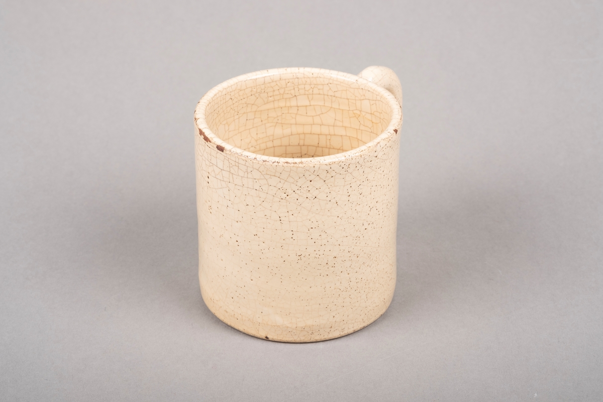 Keramikkopp med hank. Koppen er lakkert med beigefarget glasur. Det er krakeleringer på på hele koppen.