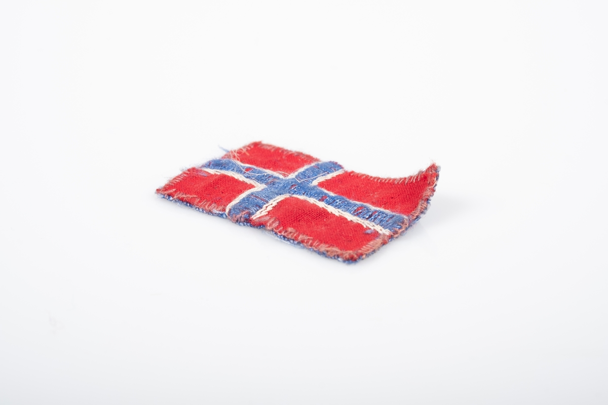 Et lite norsk flagg. Den er sydd og brodert på hånd. Den er sydd sammen av to stoffbiter, rød på forsiden og blå på baksiden. Det blå og hvite krysset på flagget er brodert.