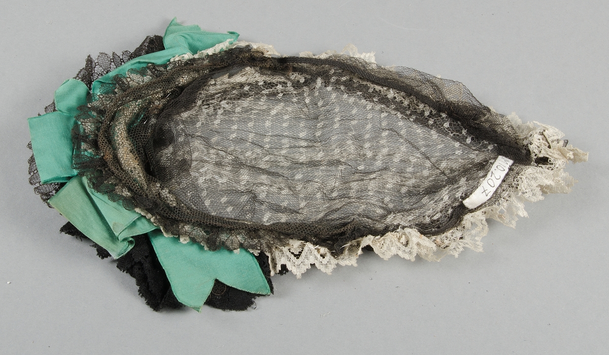 Hårklädsel av svart och vit tyll med spetskant. På huvudet rysch av svart bomulls-och tyllspets och gröna sidenband. Ståltrådsstomme.
