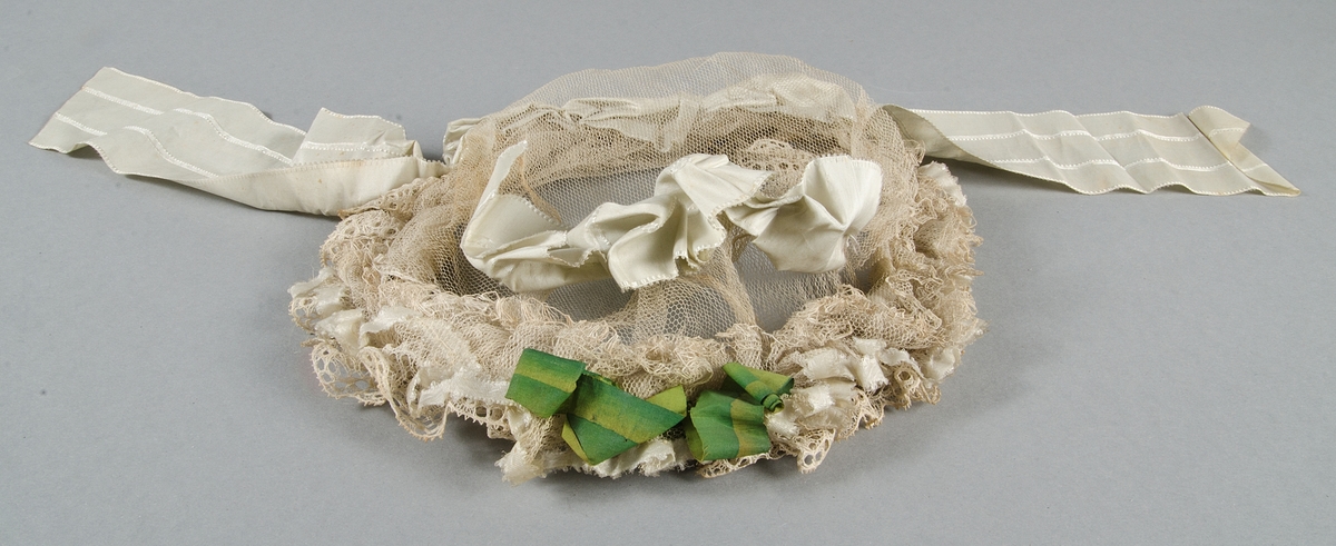 Hårklädsel avv vit tyll. Rysch av vit spets. Prydd med vaxade bomullsband i två gröna nyanser. Rosetter och band av vitt siden med mönstervävda bårder.