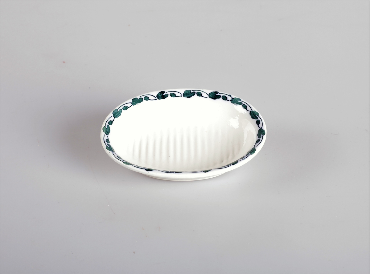 Skåla er oval med riflete bunn, og den er hvitglassert med blågrønn blomsterranke på innersiden av kanten.