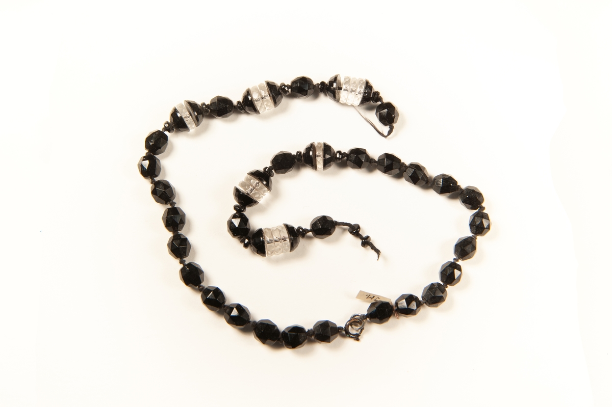 Halsband av svarta, fasettslipade stenkolspärlor med sex pärlor uppblandade med klara, slipade glasdiskar. Mitt fram är halsbandet trasigt och pärlor saknas.