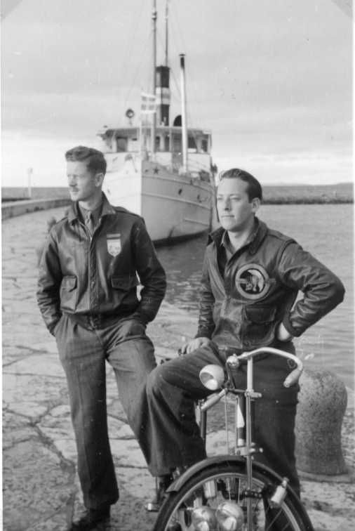 Två män i skinnjacka med emblem vid Gränna hamn, den ena sitter grensle över en cykel. I bakgrunden ligger troligen Motala Express.