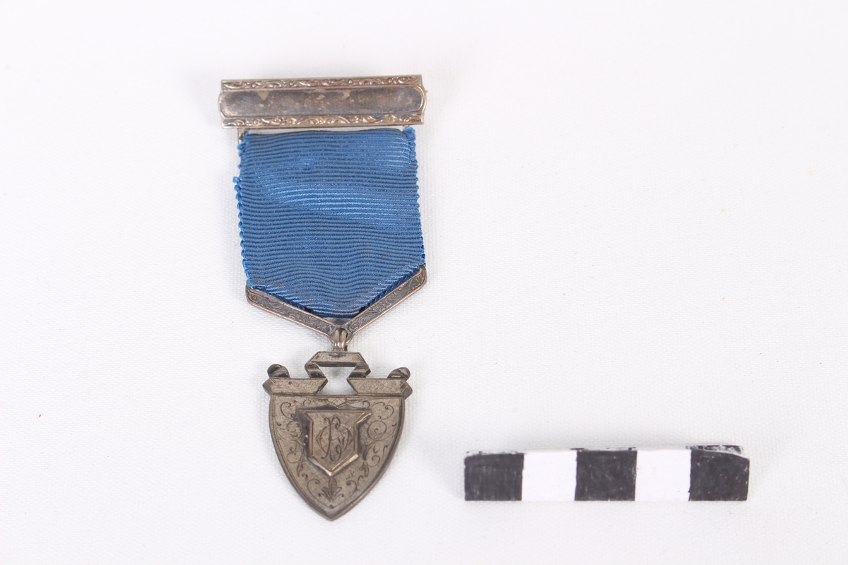 Medalje formet som et skjold med blått medaljebånd og agraff.