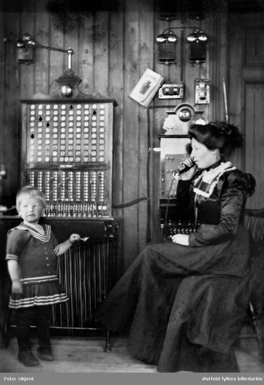 Rakkestad Telefonsentral 1902. Til høyre Mina Torper  telefonistinde ved sentralbordet. Sønnen Christoffer, tre år er med mor på jobb.