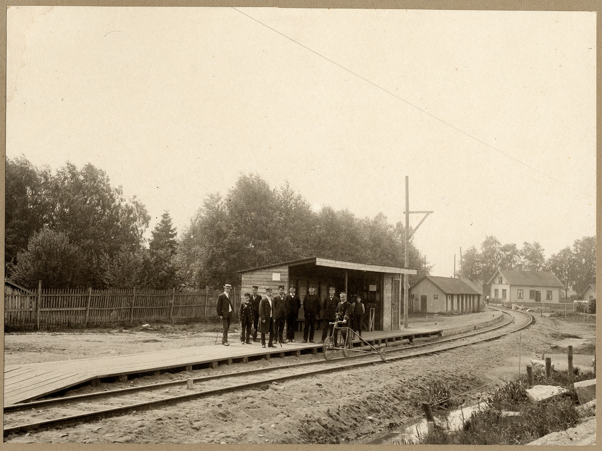 Tidaholm 1907. 10 personer framför ett skjul. Man på trehjulig trampdressin
