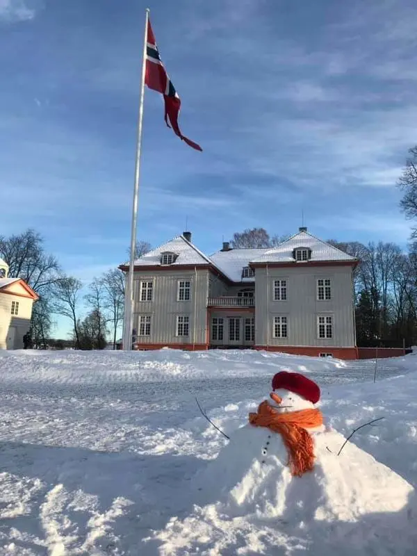 Fotografi av en liten snømann med lue og skjerf en solfylt vinterdag. På bakken snø over alt, i bakgrunnen Eidsvollsbygningen