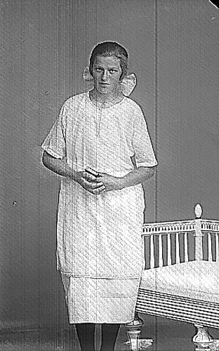 Stina Magnusson, en flicka i ljus klänning, rosett i håret och troligen psalmbok i händerna, stående intill en soffa i sengustaviansk stil. Troligen bild i samband med konfirmation.