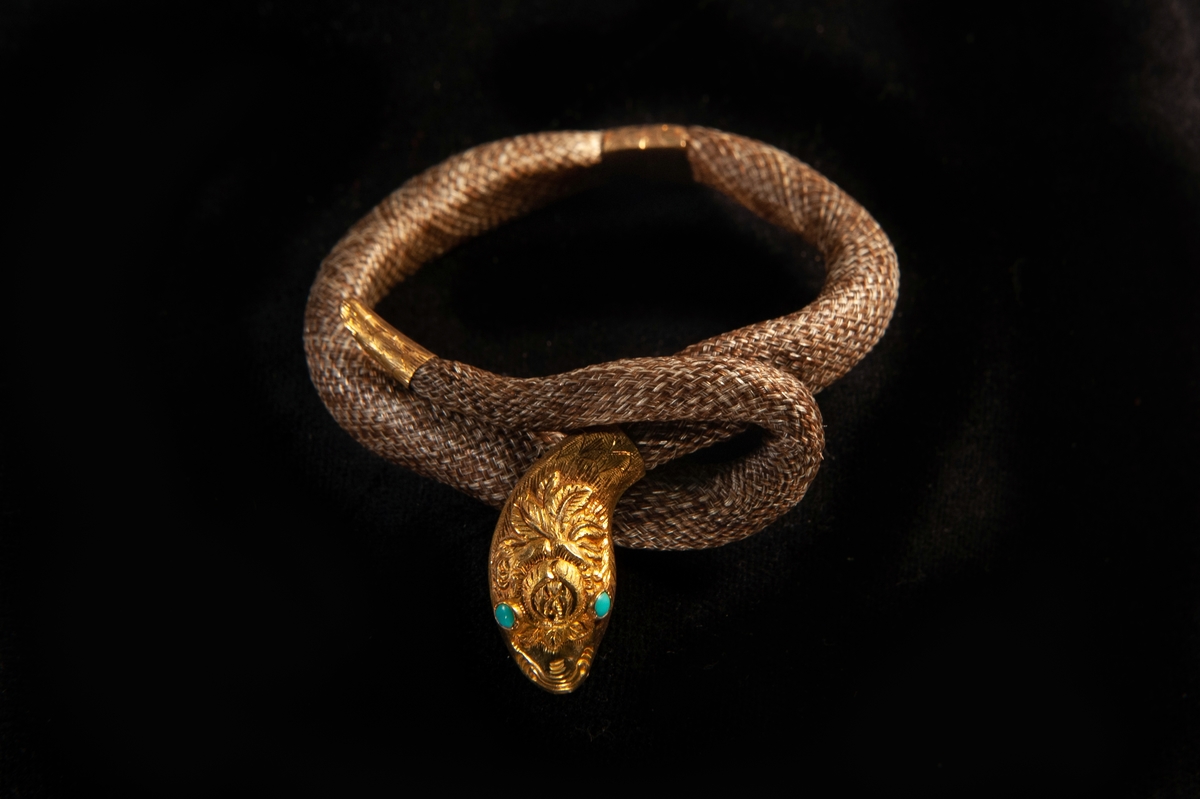 Ett armband av cendré hår i flätat hårarbete. Formad som en orm med huvud, stjärt och lås i guld. Turkoser infattade som ögon. Graverad dekor på huvudet. Stämplar bak på huvudet.