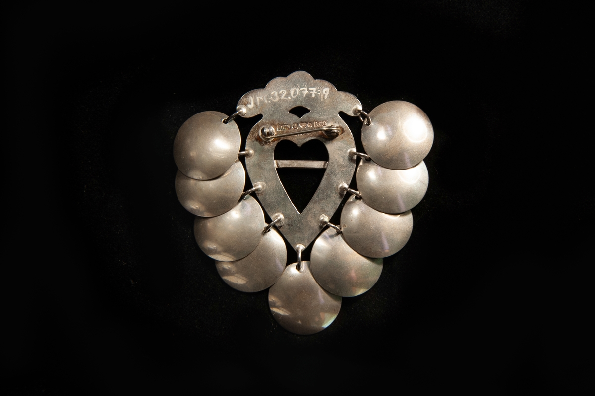 Hjärtformad sölja av silver med stiliserad krona överst, prydd med graverad blomdekor. Behängd med nio runda silverplattor ("skålar").