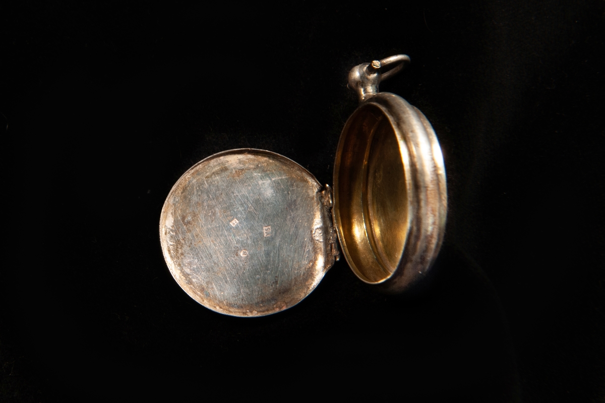 Medaljong i form av en klockboett. Stämplar invändigt på locket: . Släta sidor, en knopp upptill med en oval ring att hänga kedjan i. Invändigt förgylld.