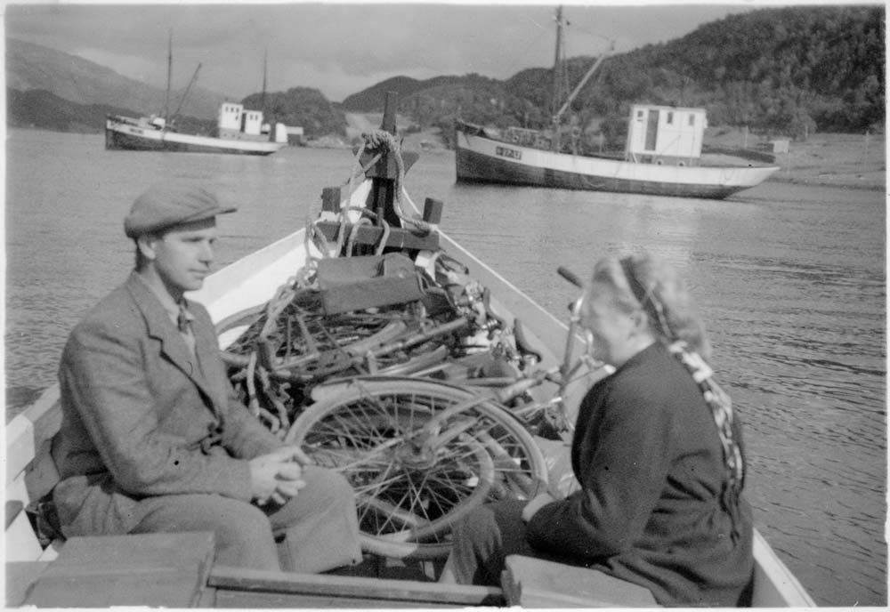 Mann og kvinne som sitter i en båt med flere sykler som ligger i den. Flere fiskebåter på sjøen.