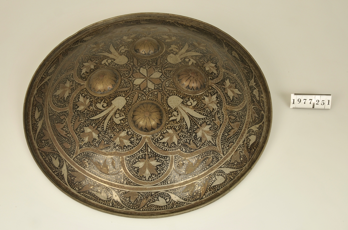
Sköld tillverkad av försilvrad koppar med rikt dekorerad framsida. Rund form, orientaliskt arbete.