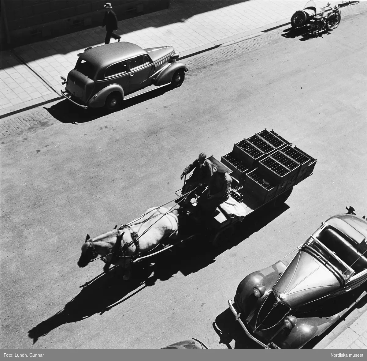 Döbelnsgatan, Stockholm, fotograferad från Gunnar Lundhs bostad. Hästvagn från AB Stockholmsbryggerier lastad med ölbackar kör på gatan. På andra sidan gatan en Chevrolet och en Harley Davidson. Närmast i bild en Studebaker av 1934 års modell.