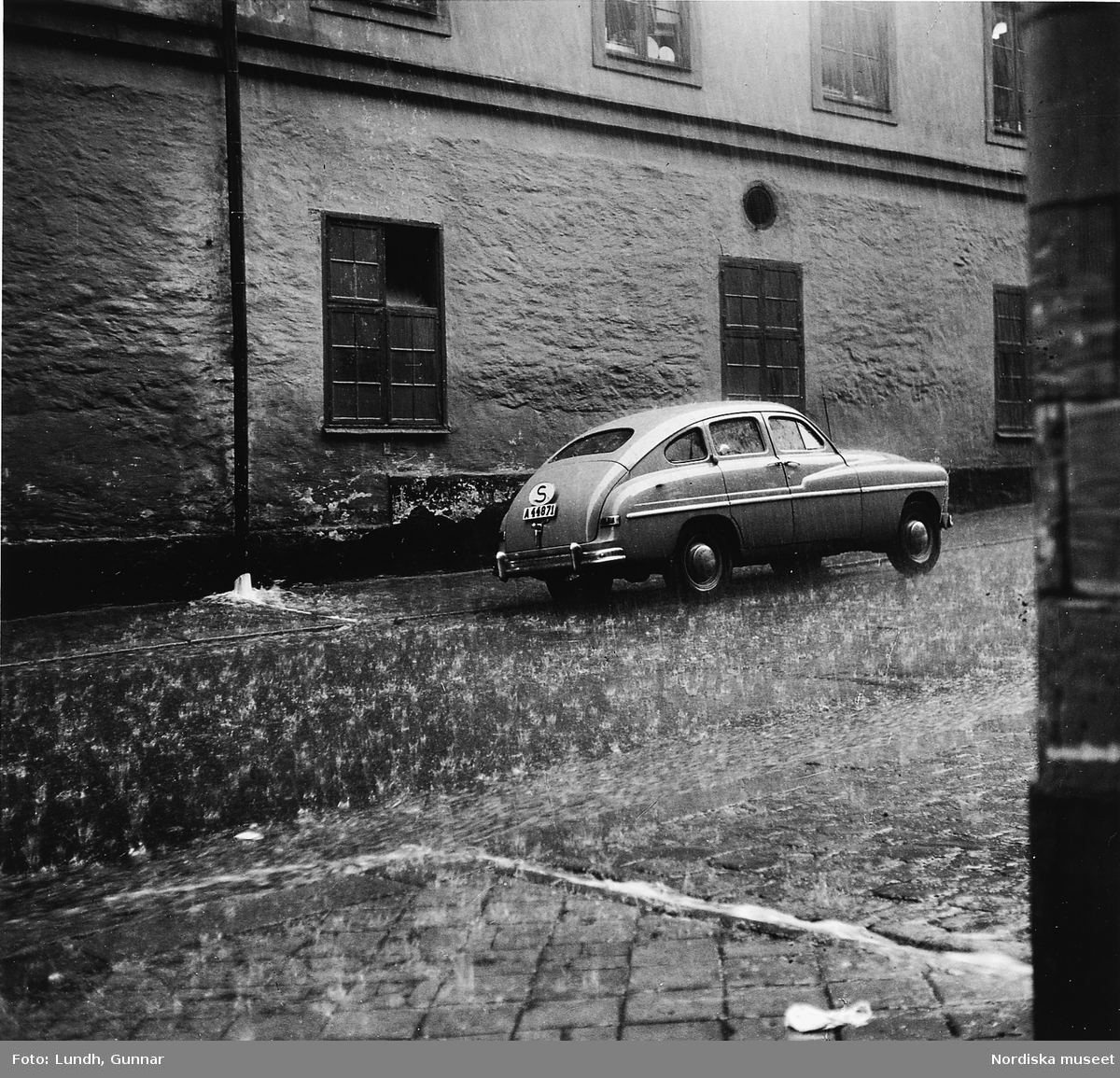 Kraftigt regn på en stadsgata, en Ford Vedette står parkerad.