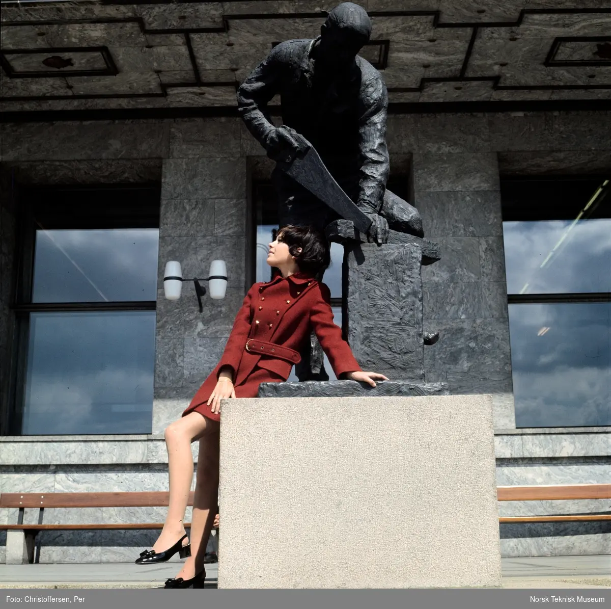 Motebilde i farger. Kvinnelig modell i rød ullkåpe, sittende på statuen "Snekker" (1954) av Per Palle Storm foran Rådhuset i Oslo, 1968. Bildet er en del av en serie der noen er publisert i ukebladet Det Nye samme høst.