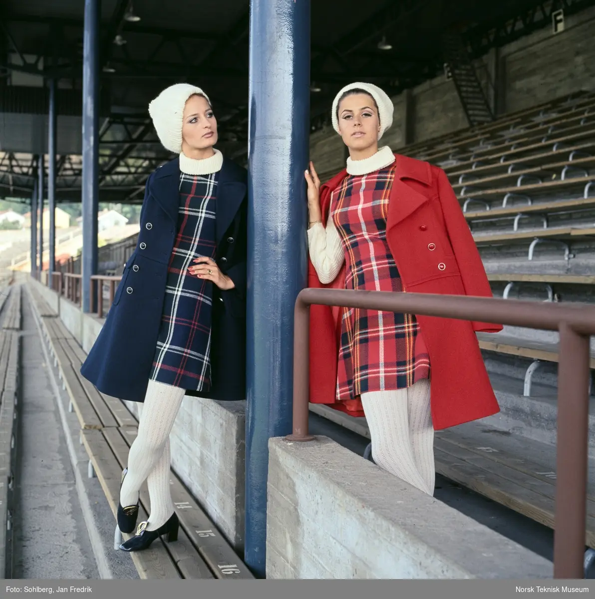 Motebilde i farger. To kvinnelige modeller i blå og røde antrekk med matchende kåper og skotskrutete kjoler, hvite strømper og hvite luer. Poserer på tribune på idrettsplass. Motivet er en del av en serie der noen er publisert i ukebladet Det Nye 1969, nr. 38 med overskriften "Kåper i høst".