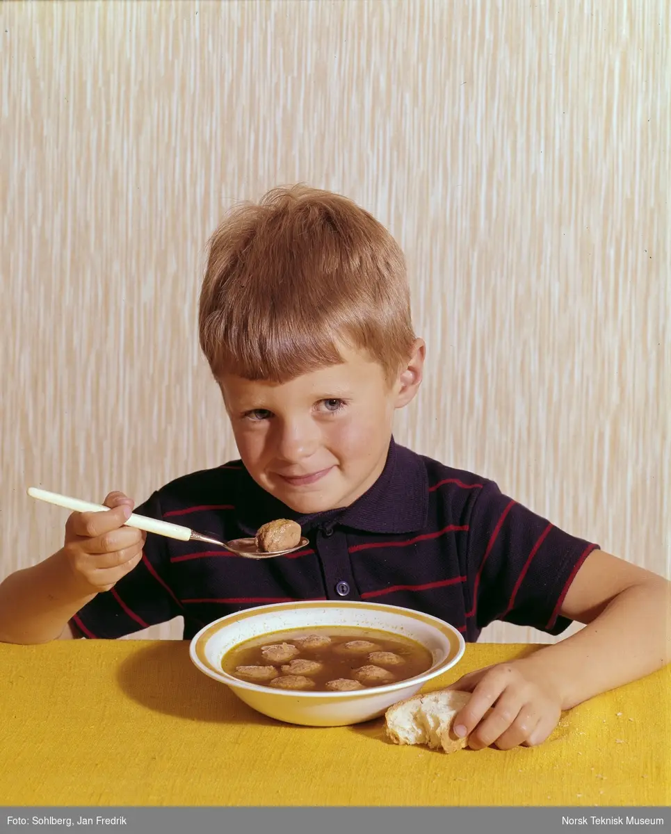 Reklamebilde for kjøttboller av typen Bolls. Hermetisk norsk bondekost. En liten gutt smiler mens han spiser kjøttboller og saus.