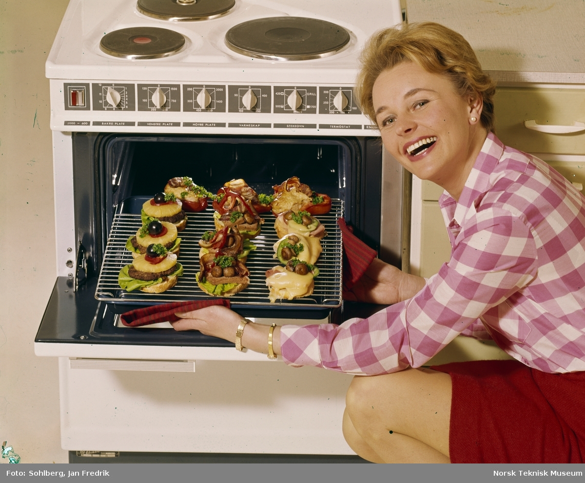 Reklamefoto for komfyr. Med moderne kjøkkenutstyr er livet som husmor ganske enkelt. En smilende kvinnelig modell lager lune smørbrød i stekeovnen.