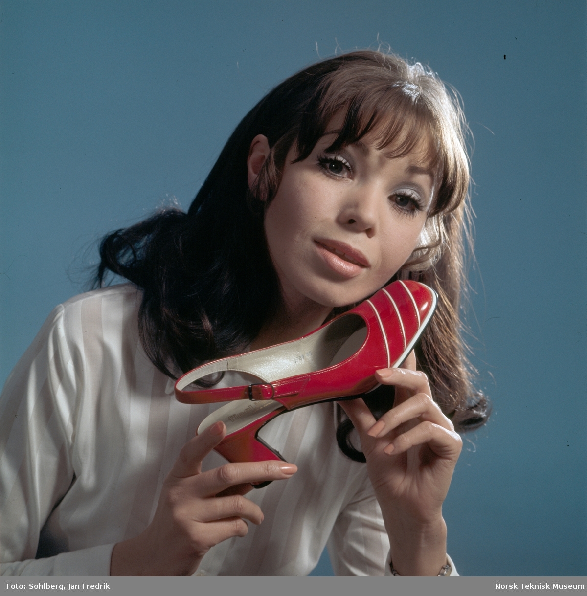 Tidlig norsk reklame- / motefoto. En kvinnelig modell holder en rød sko med hvite striper opp mot kinnet. Hun virker fornøyd med tidens skomote.