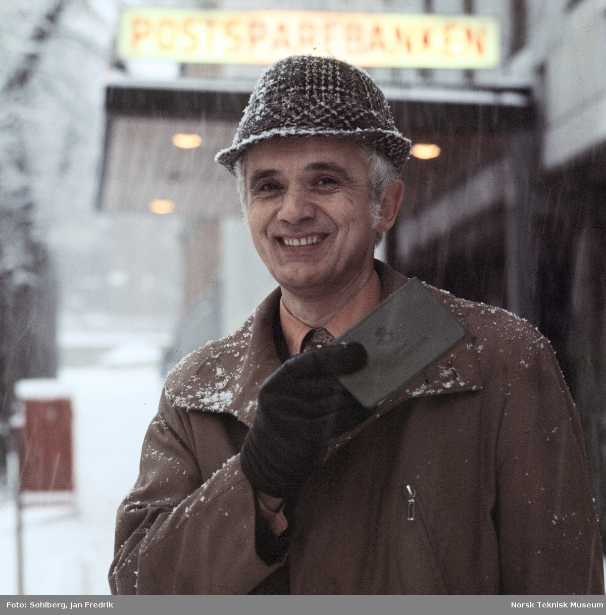 Mann med hatt og frakk utenfor Postsparebanken, holder sjekkhefte, trolig reklame for Postsparebanken. Usikker datering.
