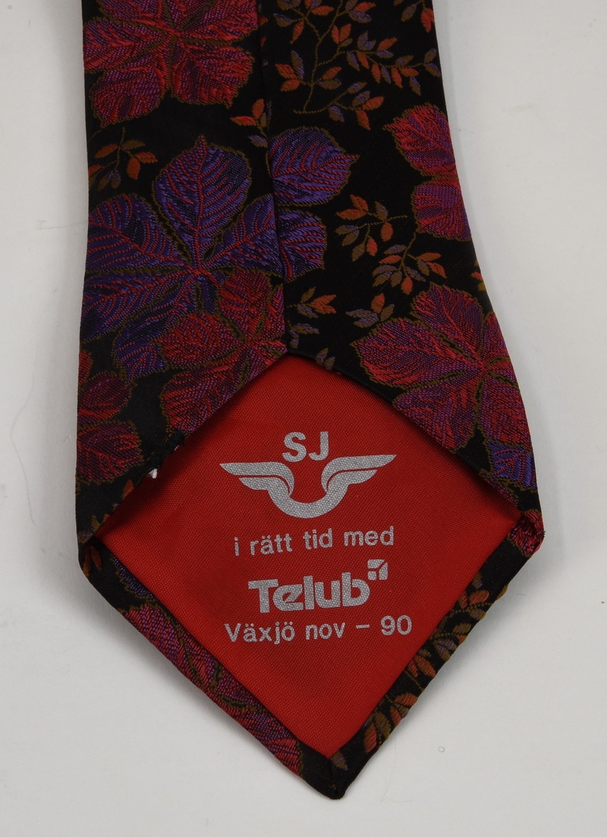 Mönstrad slips med svart botten. Mönstret består av broderade blommor och lövverk i bland annat violett och rosa, mönstret upprepas längs hela slipsen. Slipsen är fodrad med en röd textil med grå text.