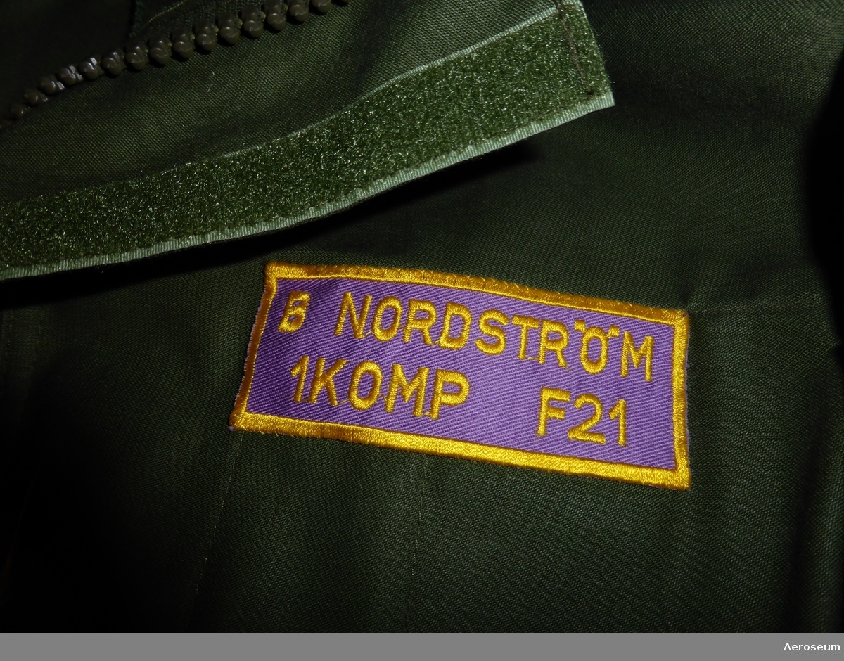 En mörkgrön overall för färdmekaniker. Denna har använts i svenska flygvapnet och av B. Nordström. Det finns på overallen en namnskylt där det står: "B NORDSTRÖM 1 KOMP F 21". På ena armen finns det ett förbandsmärke där det står "1 KOMP [bild på ett flygplan, troligtvis Viggen] F 21/SeÖN". På andra armen sitter ett vapenslagsmärke som föreställer det svenska flygvapnets.