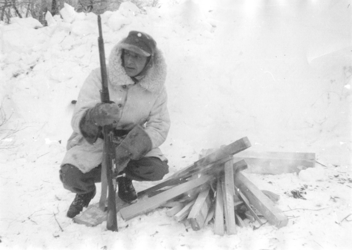 Lägereldsvakt vid Svenska frivilligkåren i Finland, F 19, 1940. En man i vinteruniform och med gevär.