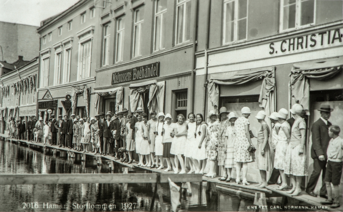 Postkort, Hamar, Strandgata, Flommen 1927, Mjøsflommen 1927, plankefortau med mye folk oppstilt foran Magnussens Bokhandel. Klokkehatter. 