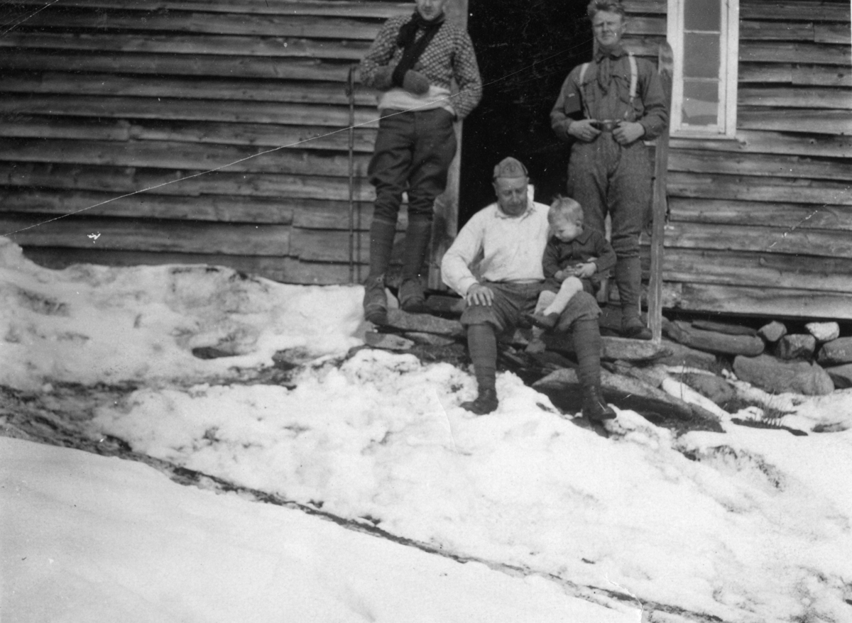 Tre menn og en liten gutt foran en bygning på Fjellberg ved Ulladalen i Suldal. Fotografens bror, Einar, har armen i fatle. Faren, Paul, sitter på trappa og Rasmus Kyvik står til høyre.