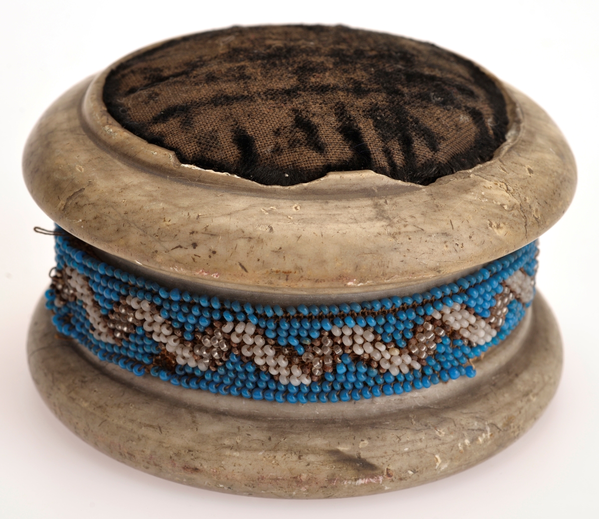 Rundformet kleberstein med nålepute av sort fløyel på toppen. Pyntet med mønstret perlebånd med hvite, blå og gjennomsiktige glassperler på midten. Perlene er sydd på stramei.