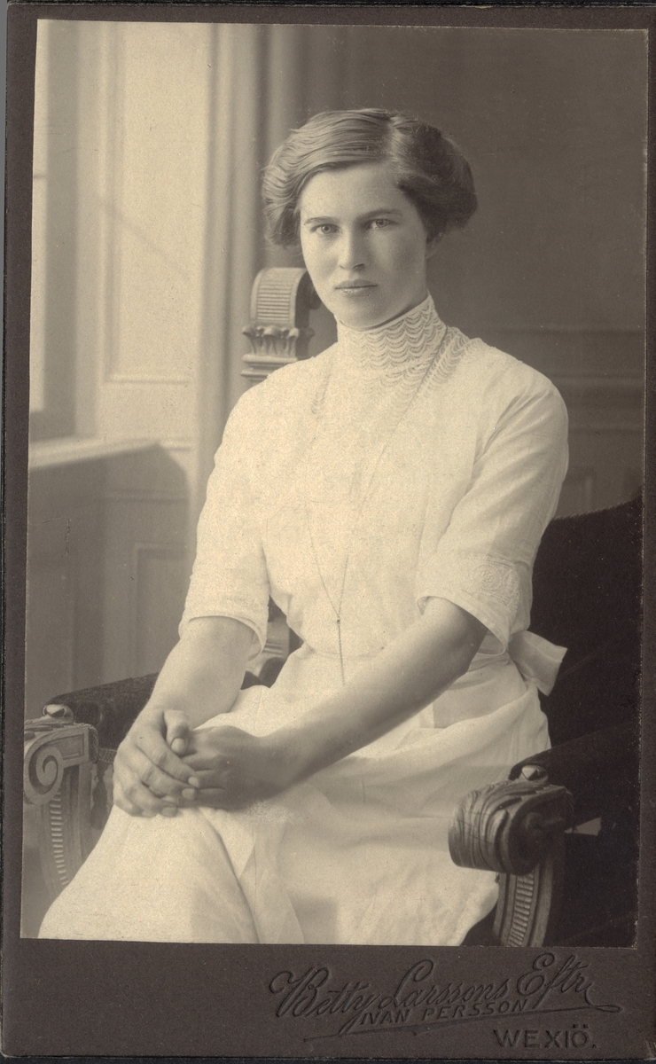 Porträttfoto av en ung, sittande kvinna i tvådelad ljus klänning med hög krage och diverse spetsbroderier på krage och blus.
Knäbild, ateljéfoto.