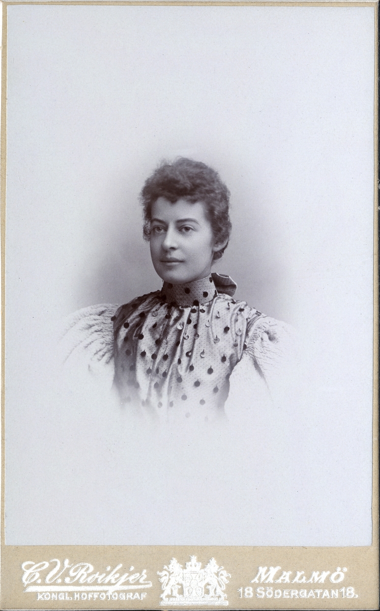 Porträttfoto av en okänd kvinna klädd i höghalsad blus med paljetter. 
Bröstbild, halvprofil. Ateljéfoto.