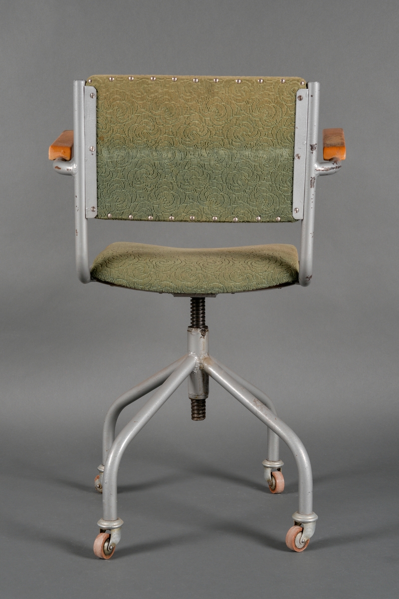 En kontorstol med fire hjul. Armlenene er laget av heltre og er lakkert (blank lakk) mens sete og rygg er laget av kryssfinér. Både sete og rygg er polstret og trukket med tekstil. Tekstilet er grønt og har vevd mønster i seg. Stolen er festet på en stålrørkonstruksjon som er grålakkert. Stolen er festet til foten med en skrueanordning og kan skrues opp og ned. Stolen ble pusset på 1960-tallet.