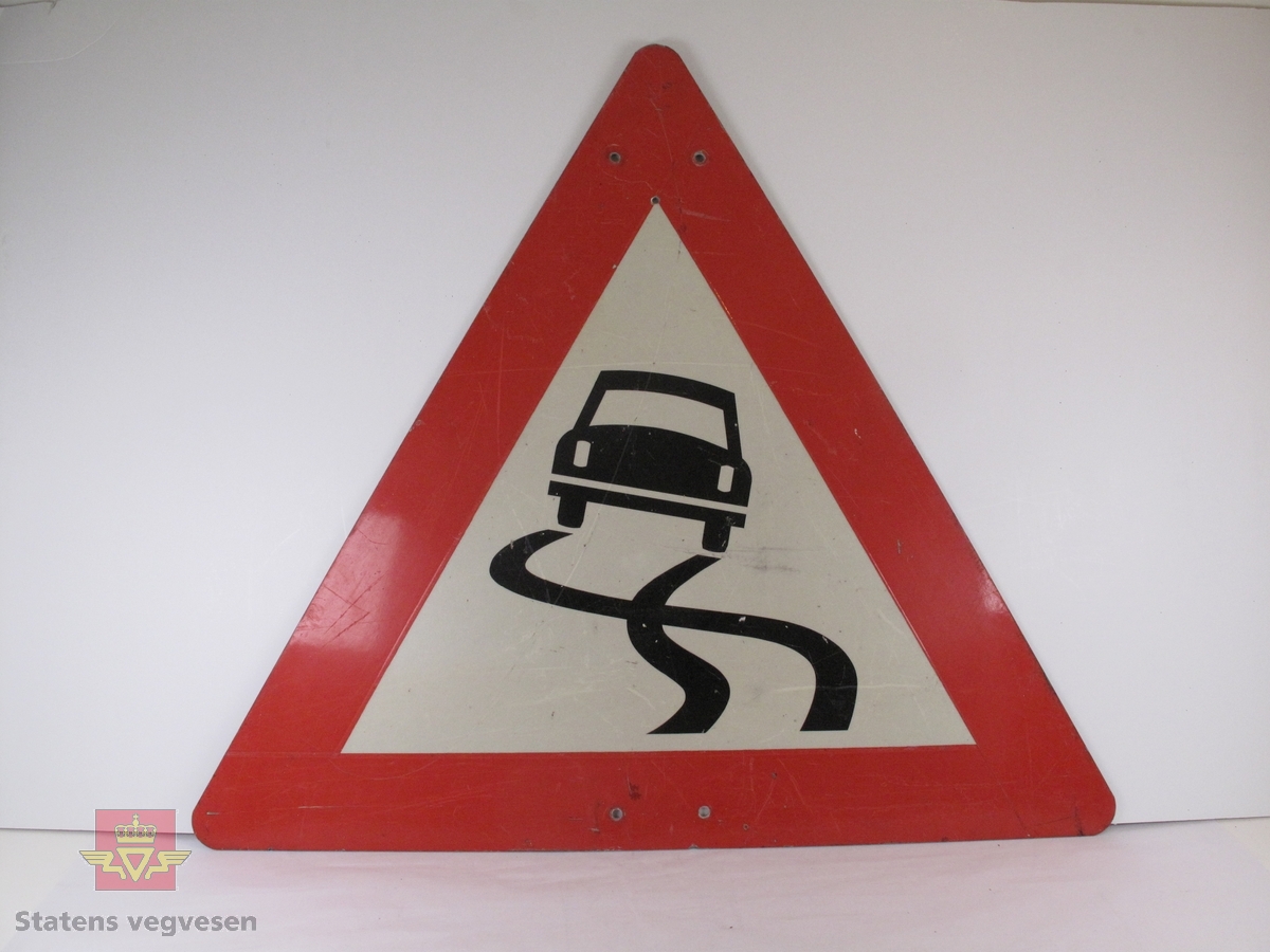 Trekantet skilt (fareskilt) med rød bord (9 cm). Figur av en bil med to bølgete streker foran, som skal illustrere glatte veger. 
Skiltet er ripet, og har noen skitne merker. Det har to skruehull på midten nede, og to helt øverst.